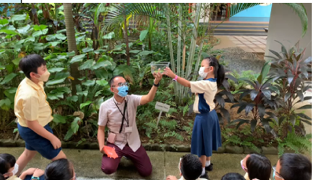 A masked teacher kneels outdoors showing a small container to a uniformed student while other children watch.
